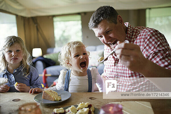 Playful father feeding cake to cute eager daughter