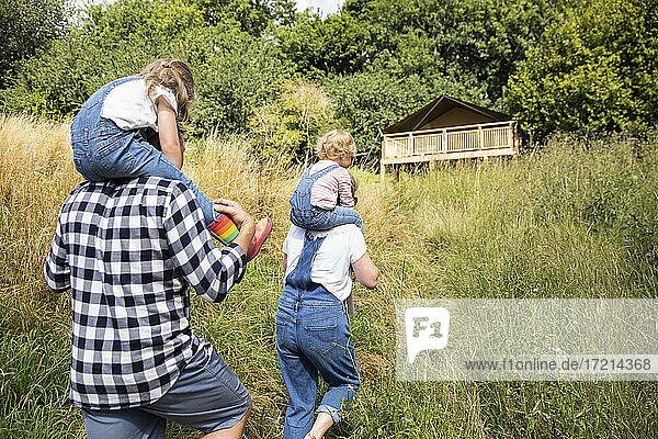 Parents carrying daughters on shoulders outside cabin in sunny woods