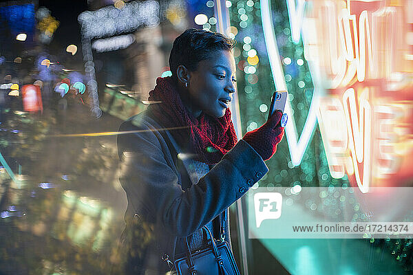 Young woman with smart phone at neon storefront sign at night