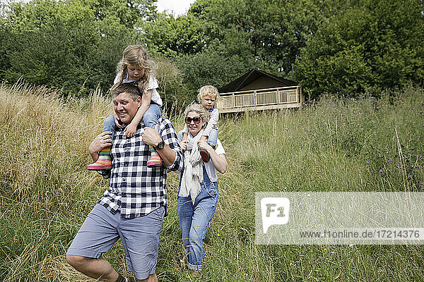 Happy parents carrying daughters on shoulder in tall grass below cabin