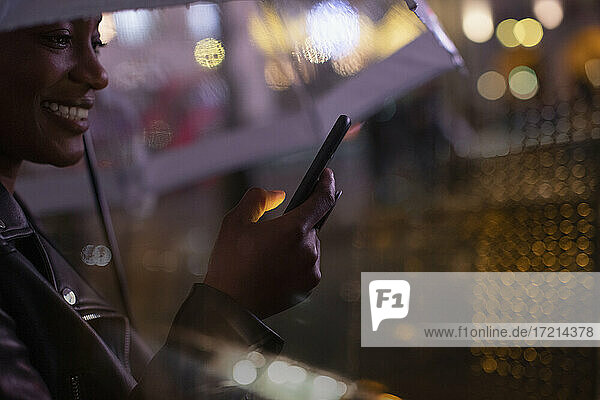 Junge Frau mit Smartphone mit Regenschirm in der Nacht