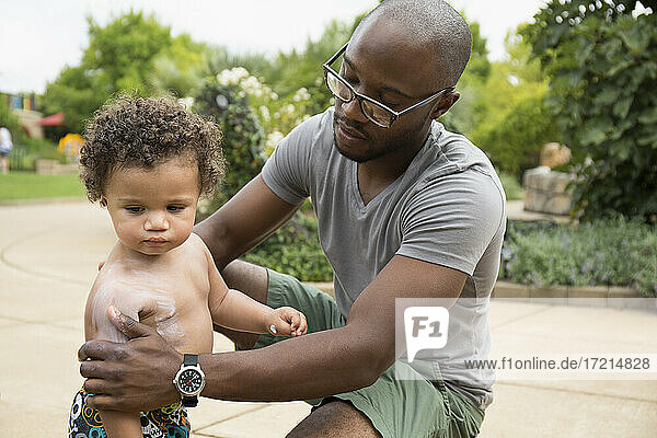 Father putting sunscreen on toddler son in park