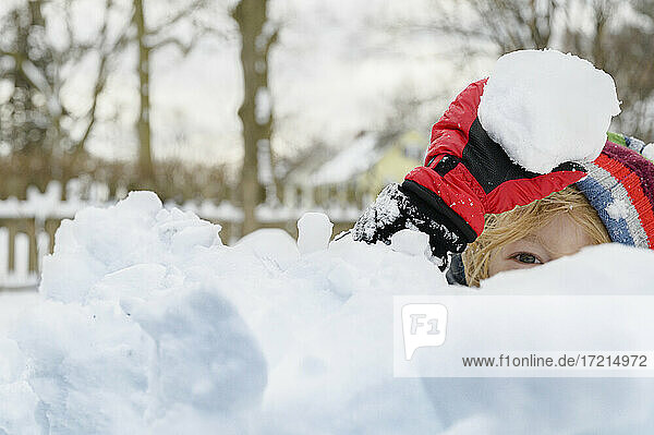 Junge (6-7) versteckt sich hinter einem Schneehaufen und hält einen Schneeball