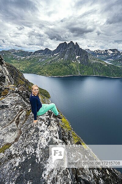 Junge Wanderin sitzt an Felsklippe  Abgrund zum Fjord  Ausblick vom Berg Barden  Senja  Norwegen  Europa