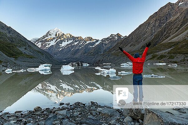 Hiker  young man standing on a rock and stretching his arms in the air  Mount Cook in morning light  Hooker Lake with reflection  sunrise  Mount Cook National Park  Southern Alps  Hooker Valley  Canterbury  South Island  New Zealand  Oceania