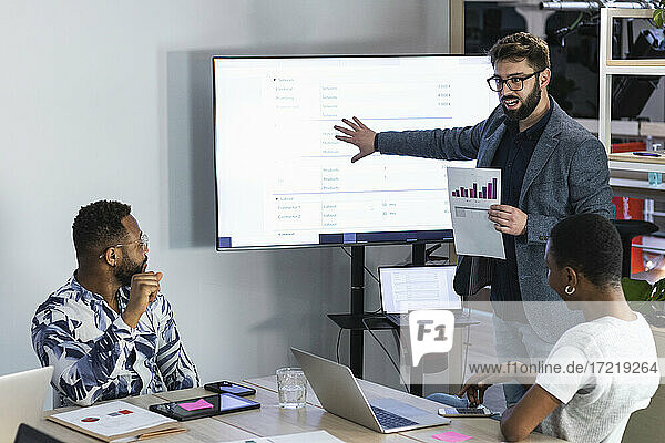 Male entrepreneur explaining data over document and television to colleague in board room