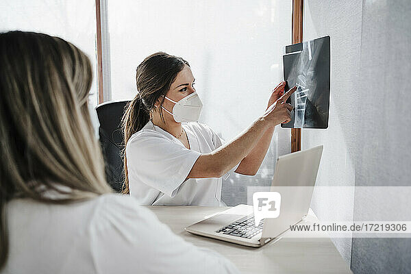 Female doctor wearing protective face mask showing x-ray to patient at desk in clinic