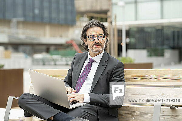 Thoughtful male professional using laptop while sitting on bench at office park
