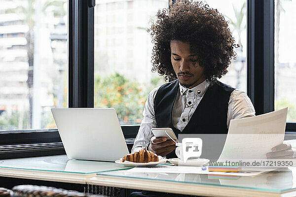 Junger Geschäftsmann benutzt sein Smartphone und hält ein Dokument in einem Café