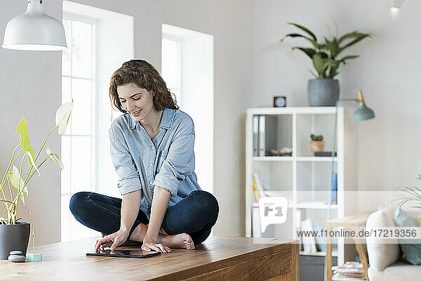 Junge Frau benutzt ein digitales Tablet  während sie zu Hause auf dem Tisch sitzt