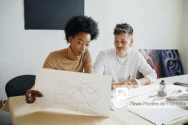 Junge Frau betrachtet kalligrafische Schrift auf Papier mit einem Mann  der an einem Tisch im Atelier sitzt