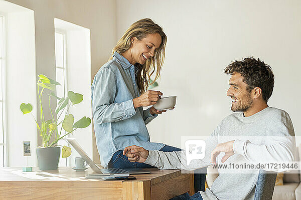 Lächelnder Freund mit Laptop  der mit seiner Freundin diskutiert  die auf dem Schreibtisch im Büro zu Hause sitzt