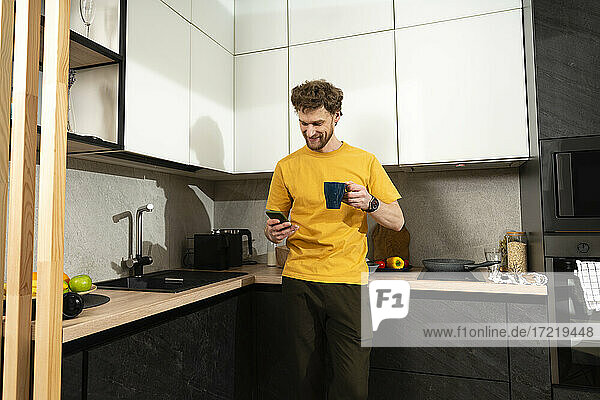 Mittlerer erwachsener Mann  der ein Mobiltelefon benutzt  während er in der Küche zu Hause Kaffee trinkt