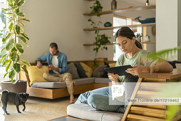 Junge Frau  die ein digitales Tablet benutzt  während ihr Freund zu Hause im Hintergrund ein Smartphone benutzt