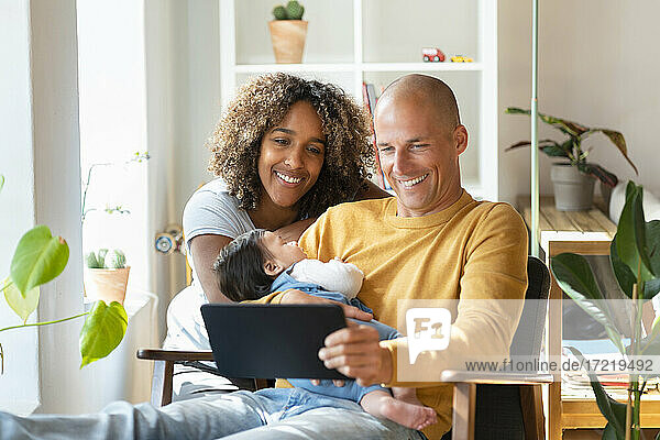 Lächelnde Eltern schauen auf ein digitales Tablet  während sie ein schlafendes Mädchen zu Hause halten