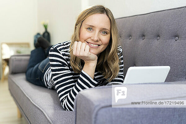 Smiling blond woman with digital tablet lying on sofa at home