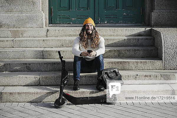 Lachender männlicher Hipster mit Mobiltelefon auf einer Treppe sitzend an einem sonnigen Tag
