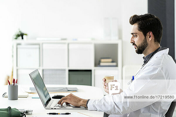 Männlicher Arzt  der einen Laptop benutzt  während er am Schreibtisch im Krankenhaus sitzt