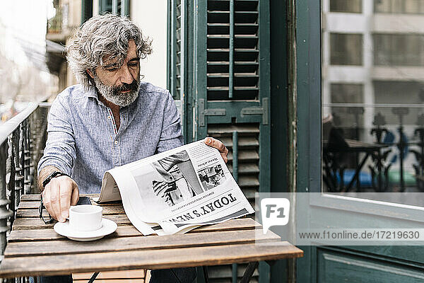 Mann trinkt Kaffee und liest Zeitung  während er am Tisch auf dem Balkon eines Hauses sitzt