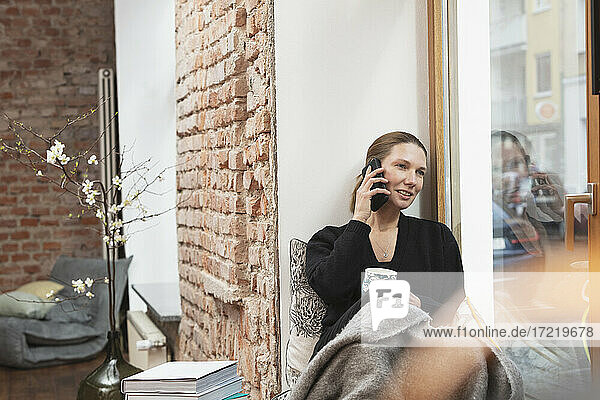 Lächelnde Geschäftsfrau mit Teetasse  die wegschaut  während sie am Fenster mit dem Handy telefoniert