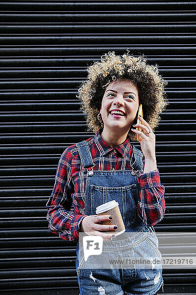 Glückliche Frau  die eine Einweg-Kaffeetasse hält  während sie mit einem Smartphone vor einem Fensterladen spricht