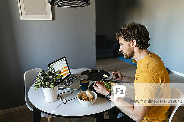 Männlicher Freiberufler beim Essen während der Arbeit am Laptop zu Hause