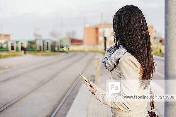 Frau mit langen Haaren  die ihr Smartphone hält  während sie am Bahnsteig auf einen Zug wartet
