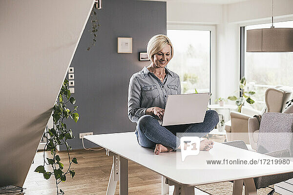Lächelnde Geschäftsfrau  die an einem Laptop arbeitet  während sie auf einem Tisch im Heimbüro sitzt