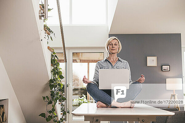 Female entrepreneur with laptop sitting in lotus position on table at home