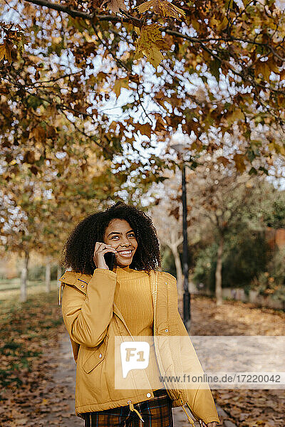Happy Afro woman talking on smart phone in public park during autumn
