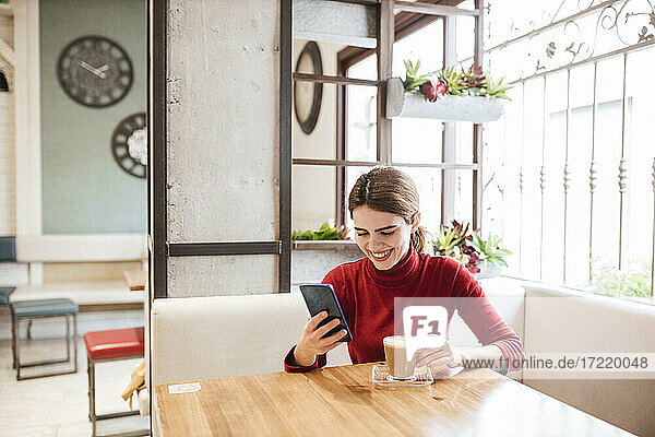 Lächelnde Frau  die ein Mobiltelefon benutzt  während sie einen Kaffee im Café trinkt