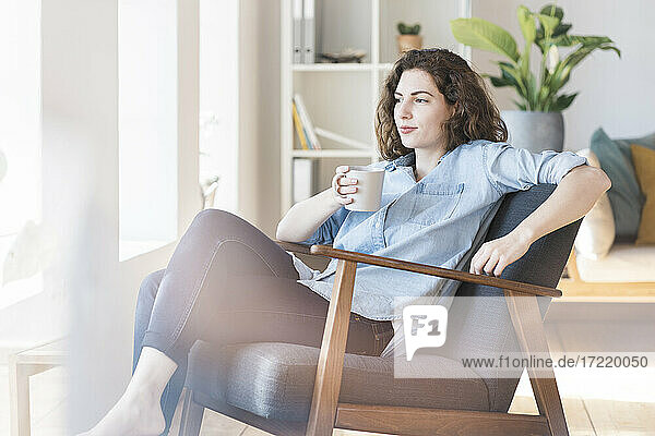 Nachdenkliche Frau  die auf einem Sessel im Wohnzimmer sitzt und Kaffee trinkt