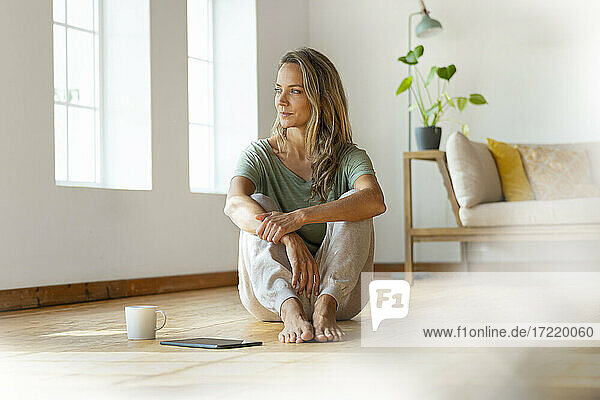 Blonde Frau träumt  während sie mit digitalem Tablet und Kaffeetasse auf dem Boden im Wohnzimmer sitzt