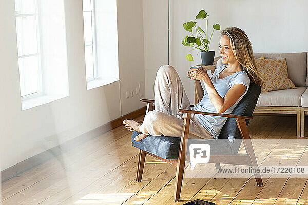 Smiling woman holding coffee mug while sitting on armchair at home