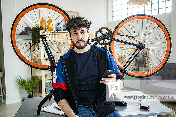 Mann hält Smartphone  während er sich zu Hause auf einen Tisch mit umgestürztem Fahrrad stützt