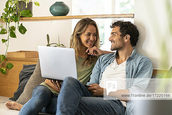 Smiling man with laptop looking at girlfriend while sitting on couch at home