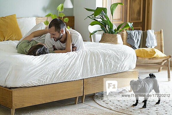 Happy young couple embracing on bed while Pug dog standing on floor at home