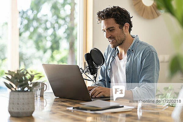 Male freelancer speaking on microphone in front of laptop while recording podcast at home