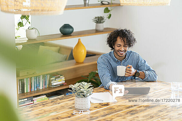 Lächelnder männlicher Unternehmer  der ein digitales Tablet benutzt  während er zu Hause Kaffee trinkt
