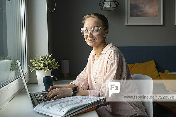 Lächelnde Frau mit Brille sitzt zu Hause am Laptop