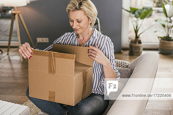 Lächelnde reife Frau  die ein Paket öffnet  während sie zu Hause auf dem Sofa sitzt