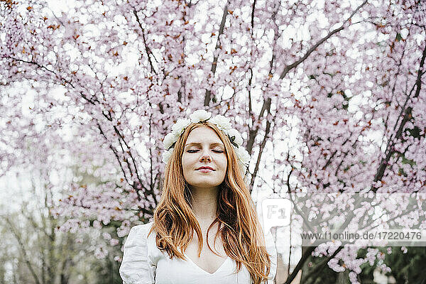 Junge Frau mit geschlossenen Augen und weißem Blumen-Diadem vor einem Mandelbaum stehend
