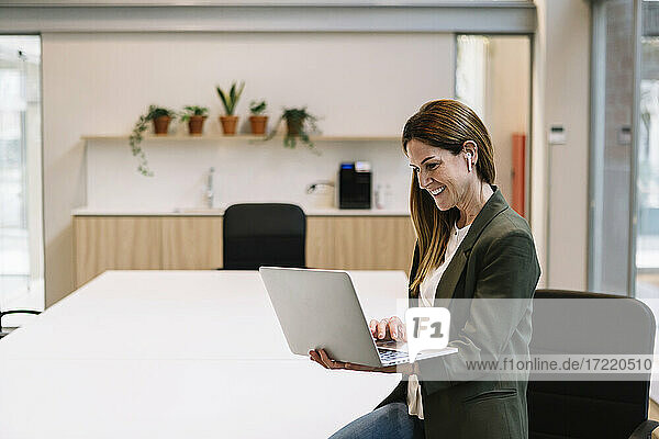 Lächelnde Unternehmerin mit In-Ear-Kopfhörern  die einen Laptop benutzt und sich auf einen Tisch im Büro stützt