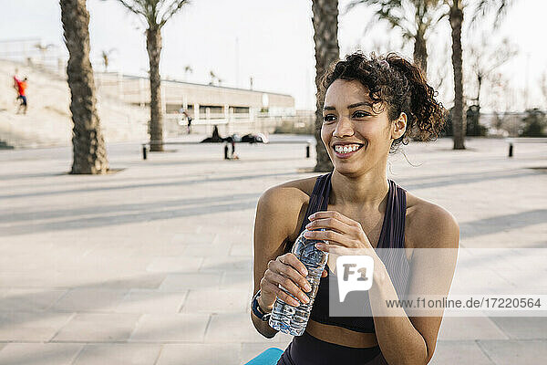 Smiling sportswoman looking away while holding water bottle on footpath