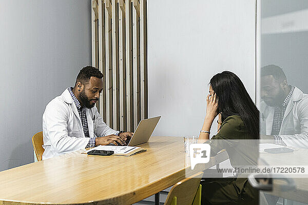 Männlicher Mediziner benutzt einen Laptop  während er mit einer Patientin in der Klinik sitzt