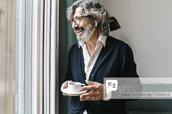 Happy senior man with in-ear headphones holding coffee cup near window at home