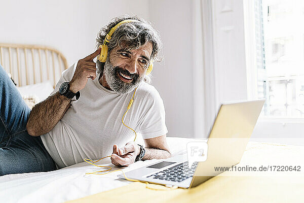 Mann berührt Kopfhörer während Videogespräch über Laptop auf dem Bett zu Hause