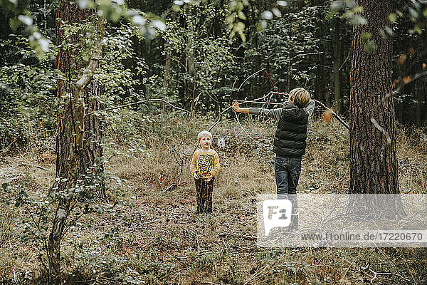 Boy playing with bow and arrow while brother standing by him in forest