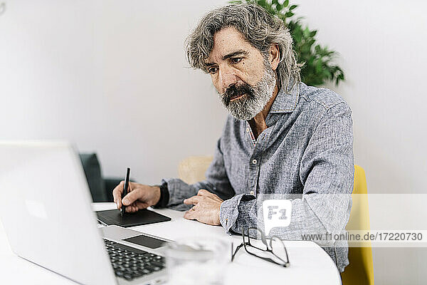 Älterer männlicher Geschäftsmann  der auf seinen Laptop schaut  während er im Büro zu Hause ein Grafiktablett benutzt