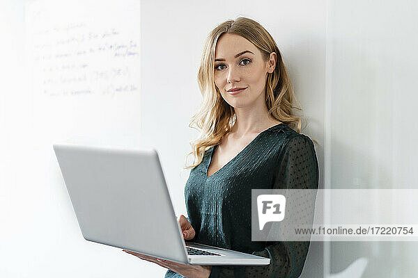 Weibliche Fachkraft mit Laptop an der Wand lehnend im Büro